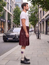 A man stands on a city sidewalk wearing a dark burgundy leather kilt, a white t-shirt, white socks, and black leather oxford shoes. A black Mercedes-Benz car is parked on the street behind him, with trees and storefronts in the background.