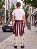 A man stands on a city sidewalk facing away from the camera, wearing an olive green pleated kilt with white vertical stripe panels, a white t-shirt, white knee-high socks, and black shoes. Storefronts, trees, and parked cars are visible in the background.