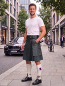 A man stands on a city sidewalk wearing a dark gray kilt, white t-shirt, white knee-high socks, and black leather shoes. The background shows a street with buildings, a parked black car, and a bicycle.