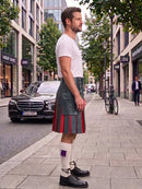 A man standing on a city sidewalk wearing a gray kilt with red and purple pleats, a white t-shirt, white socks, and black shoes, with a street scene in the background.
