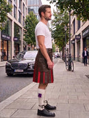 A man standing on a city sidewalk wearing a brown leather kilt with red pleated fabric, a white t-shirt, white socks, and black leather shoes. A black Mercedes-Benz car and a bicycle are parked nearby on the street.