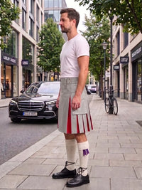 A man wearing a light gray kilt with red and purple pleated accents, a white t-shirt, white knee-high socks, and black shoes, standing on a city sidewalk next to a street with cars and trees.