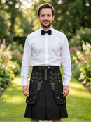 A man wearing a white dress shirt, black bow tie, and a black kilt with multiple straps, buckles, and front pockets, standing in a sunlit garden path.