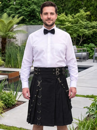 A man wearing a white dress shirt, black bow tie, and a black pleated kilt with multiple buckle and strap details, standing in a landscaped garden setting.