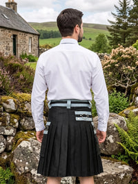 A man viewed from behind wearing a white long-sleeve dress shirt and a black kilt with a gray belt and silver buckle, standing outdoors in front of a stone wall and green hills.
