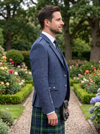A man in profile wearing a blue tweed blazer, white dress shirt, dark tie, and a green tartan kilt with a black sporran, standing in a garden path.