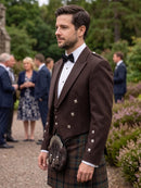 A man wearing a traditional Scottish formal outfit, featuring a dark brown double-breasted jacket with silver buttons, a white dress shirt, a black bow tie, and a tartan kilt with a sporran, standing in a garden setting with blurred guests in the background.