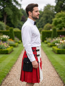 A man standing in a formal garden wearing a white long-sleeve dress shirt, black bow tie, and a red and white kilt with black belt and pouch details.
