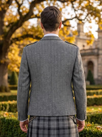 A man viewed from behind wearing a gray herringbone tweed blazer over a white collared shirt and a black and gray plaid kilt, standing outdoors in a garden with autumn trees and a stone building in the background.