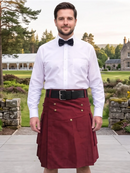 A man wearing a burgundy double-breasted kilt with gold buttons, a black belt, a white dress shirt, and a black bow tie, standing outdoors in front of a stone building and green lawn.
