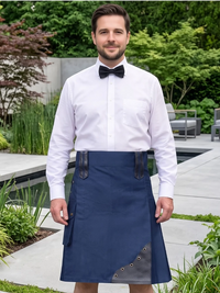 A man wearing a white dress shirt, black bow tie, and a navy blue kilt with dark leather trim, standing in a garden setting.