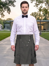 A man wearing a dark olive green pleated kilt with decorative silver buttons, paired with a white dress shirt and black bow tie, standing on a paved path in a modern outdoor setting.