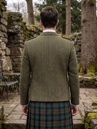 A man viewed from behind wearing an olive green tweed blazer with shoulder epaulets and a green tartan kilt, standing in a stone garden courtyard.