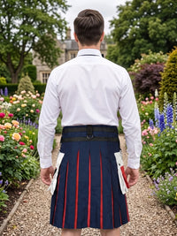 A man viewed from behind wearing a white dress shirt and a navy blue kilt with red stripes, standing in a garden path.