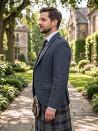 A man in profile wearing a dark navy blazer, white dress shirt, black tie, and a gray plaid kilt with a sporran, standing on a stone path in a garden courtyard with stone buildings in the background.