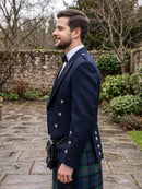A man in profile wearing a navy blue kilt jacket with silver buttons, a white shirt, a black bow tie, and a green and blue tartan kilt, standing outdoors on a stone path.