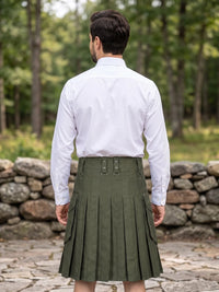 A man viewed from behind wearing a white long-sleeve button-up shirt and an olive green pleated kilt outdoors in front of a stone wall and trees.