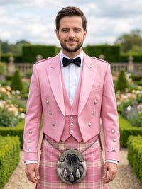 A man wearing a pink double-breasted formal jacket with silver buttons, a white shirt, a black bow tie, and a pink plaid kilt with a sporran, standing in a formal garden.