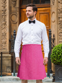 A man wearing a bright pink kilt with a white dress shirt and black bow tie, standing in front of a stone building with a wooden door.