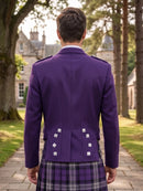A man viewed from behind wearing a purple Scottish kilt jacket with silver buttons and a purple plaid kilt, standing on a stone path in front of a large stone castle.