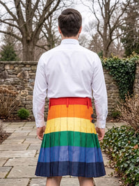 A man viewed from behind wearing a white long-sleeve button-up shirt and a pleated kilt with horizontal rainbow stripes in red, orange, yellow, green, blue, and dark navy, standing outdoors on a stone path with trees and a stone wall in the background.