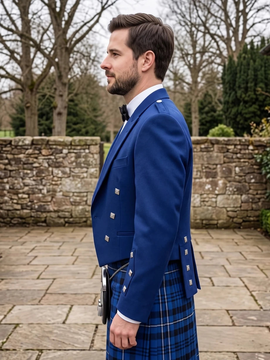 A man in profile wearing a royal blue double-breasted kilt jacket with silver buttons and a blue tartan kilt, standing outdoors in front of a stone wall and bare trees.