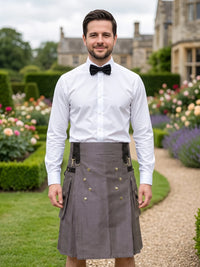 A man wearing a white dress shirt, black bow tie, and a gray kilt with black suspenders and brass buttons, standing in a garden path with a stone building in the background.