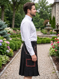 A man in a garden wearing a black kilt with red trim and a white dress shirt, shown in profile. The background features a gravel path, green hedges, and a stone building.