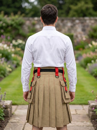 A man viewed from behind wearing a white long-sleeve shirt, a beige pleated kilt with red suspenders and side pockets, and a black belt, standing in a garden pathway.