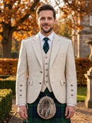 A man wearing a traditional Scottish formal outfit, featuring a light beige linen blazer and waistcoat over a white shirt, a dark green patterned tie, and a green tartan kilt with a sporran, standing outdoors in front of autumn trees.