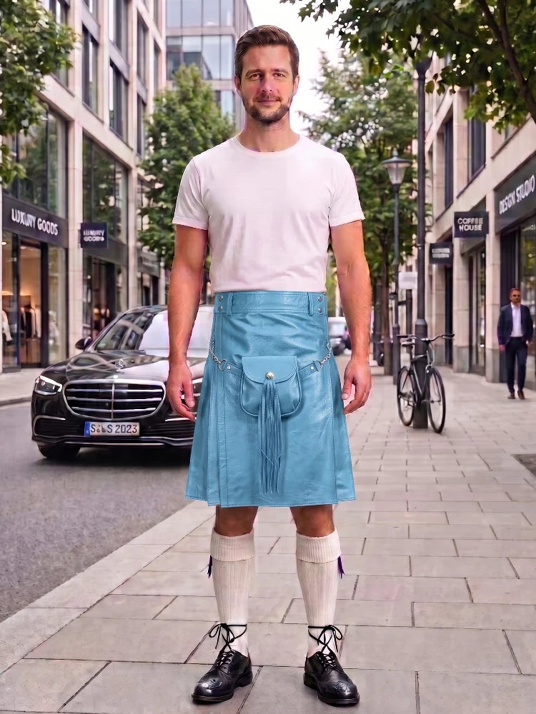 A man stands on a city sidewalk wearing a light blue denim kilt over a white t-shirt, cream knee-high socks, and black leather shoes. A black Mercedes-Benz is parked behind him on the street, with storefronts and trees in the background.