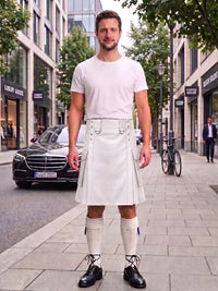 A man stands on a city sidewalk wearing a white short-sleeve t-shirt and a white kilt with belt loops, paired with white knee-high socks and black leather shoes. Buildings, trees, a parked black car, and a bicycle are visible in the background.